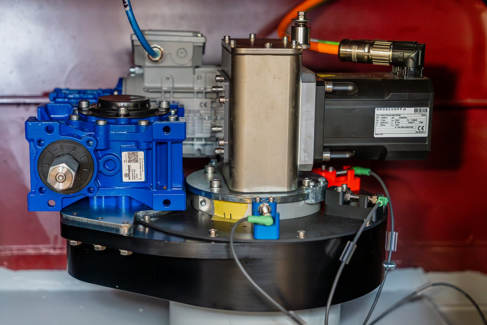 A RotorSwing drive assembly built into a hull: blue gearbox, electric motor and stainless shaft housing bolted to a black mounting plate against a red-painted bulkhead