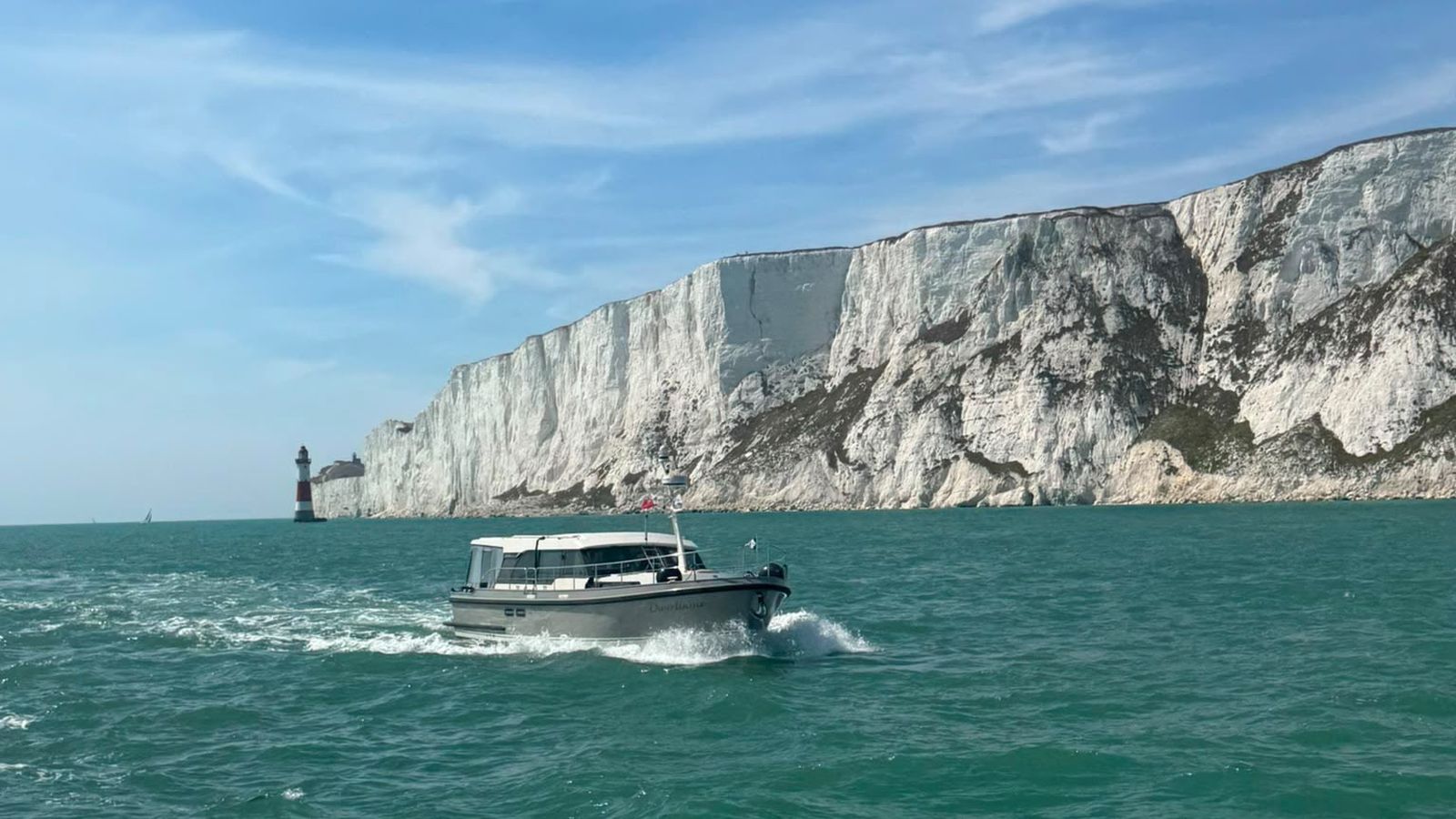 A motor yacht under way in turquoise water past white chalk cliffs, a red and white lighthouse standing at their foot