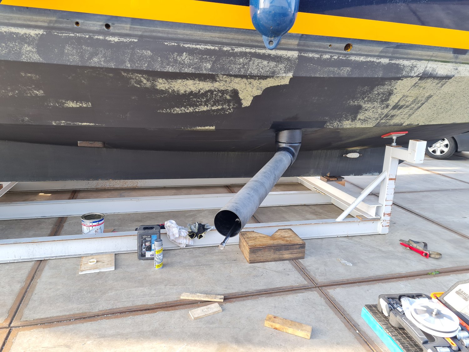 A dark blue hull standing on a boatyard cradle with a black rotor tube swung down from the hull, antifouling partly sanded back and paint tins on the concrete