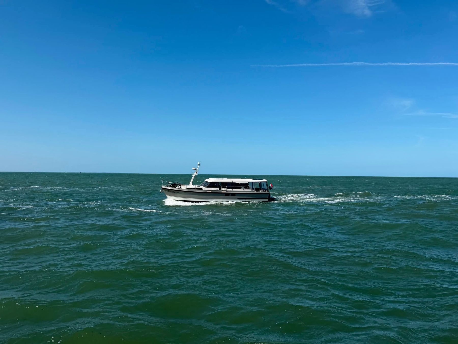 A motor yacht under way alone on open blue water under a clear sky, holding a straight line